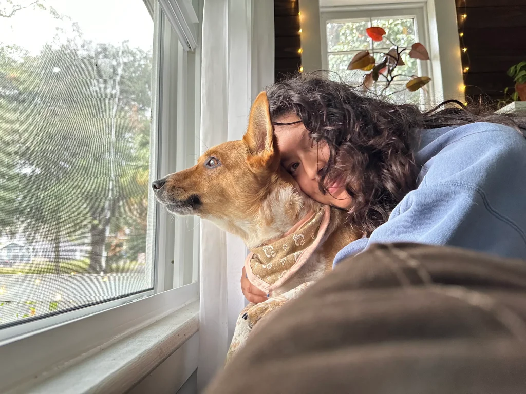 Cassandre smiling at the camera, hugging their dog who is looking out the window